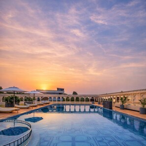 A swimming pool with a sunset view, surrounded by a covered walkway with umbrellas and a building with arches. - WelcomHeritage Akashganga Resort & Spa (Bichhiwara)