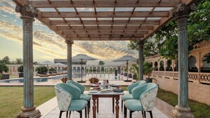 A covered outdoor dining area with a table and chairs, surrounded by columns and a view of a pool and buildings in the background. - WelcomHeritage Akashganga Resort & Spa (Bichhiwara)