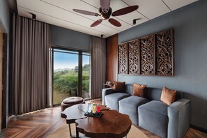 A modern living room with a large window, a ceiling fan, and a wooden coffee table with drinks. - WelcomHeritage Akashganga Resort & Spa (Bichhiwara)