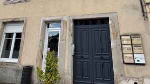 Un edificio de dos pisos con fachada color beige, varios ventanales y una puerta azul oscuro. Hay una planta en maceta frente al edificio.