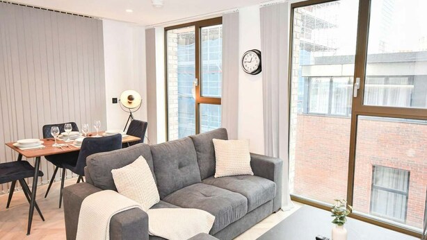 Una sala moderna con un sofá gris, una mesa de comedor con platos y vasos, y ventanas amplias con vistas a un edificio de ladrillo.