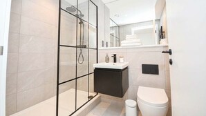Un baño moderno con ducha envuelta en vidrio, inodoro blanco, tocador negro y lavamanos blanco.