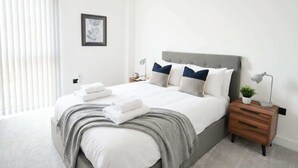 Un dormitorio moderno con cama, mesitas de noche, una lámpara y un cuadro enmarco, junto a una ventana con persianas.