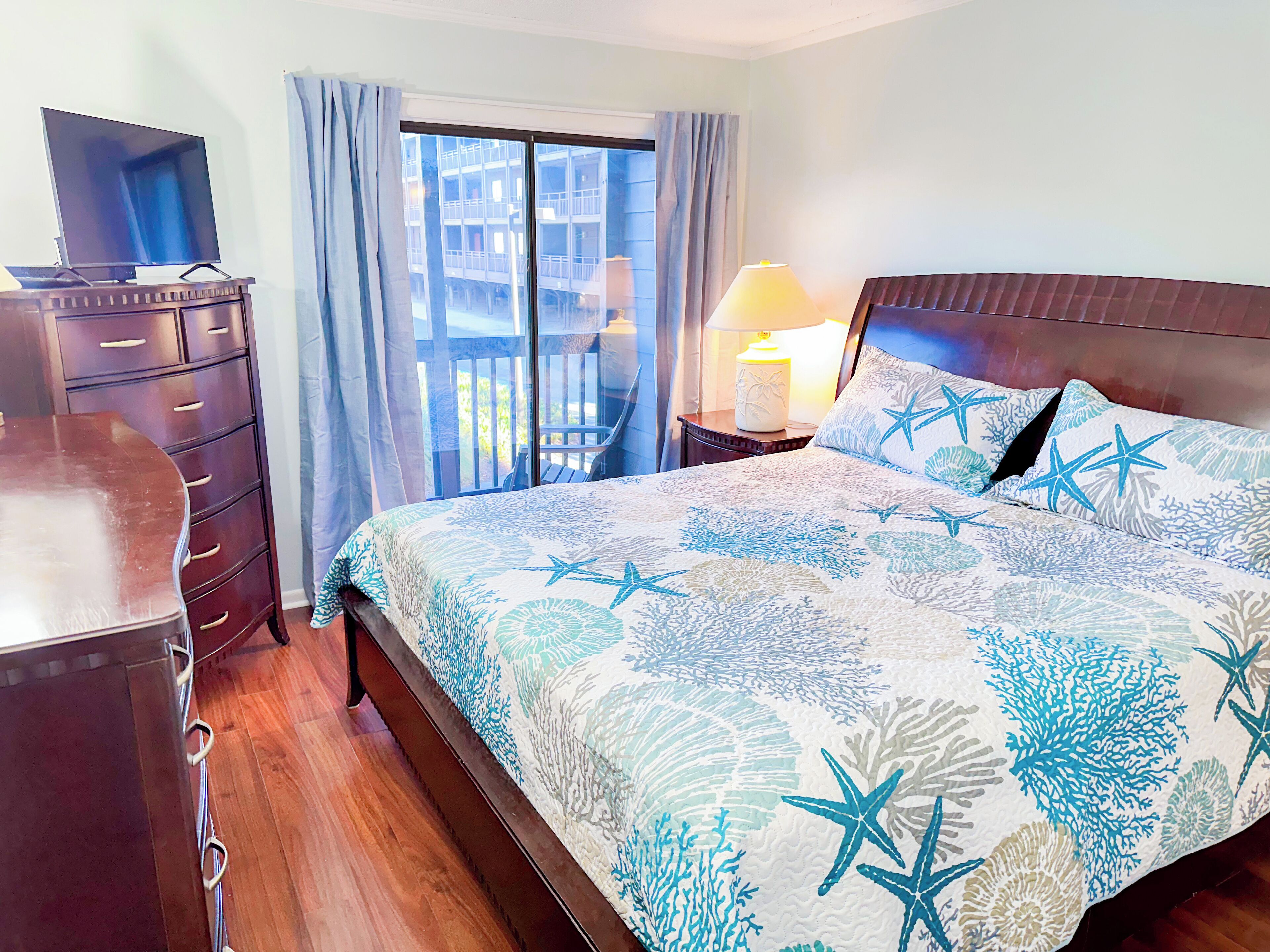Une chambre à coucher comprenant un lit, une commode, un téléviseur, une fenêtre avec des rideaux et une table de chevet avec une lampe.