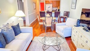 A living room with a glass coffee table, a sofa, a TV, and a dining area with chairs and a table.