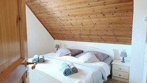 Un dormitorio con techo de madera, una cama con toallas enrolladas, una mesita de noche con lámpara y un aparador blanco.