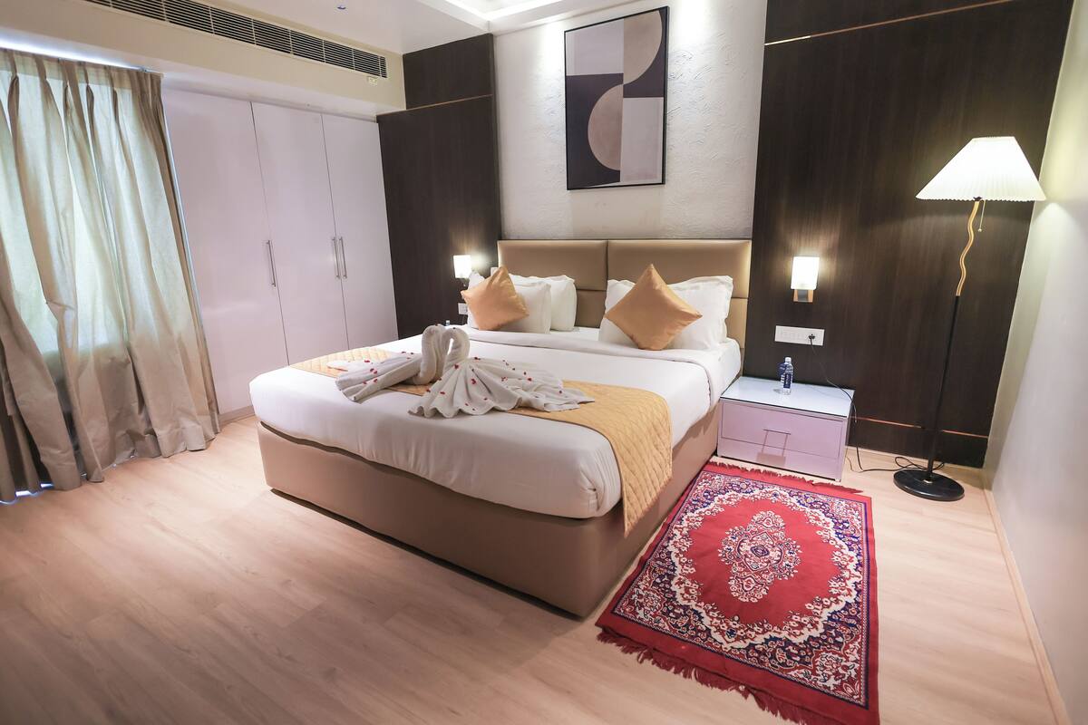 A hotel room with a bed, bedside tables, a nightstand, a lamp, a red patterned rug, and a large abstract painting on the wall.