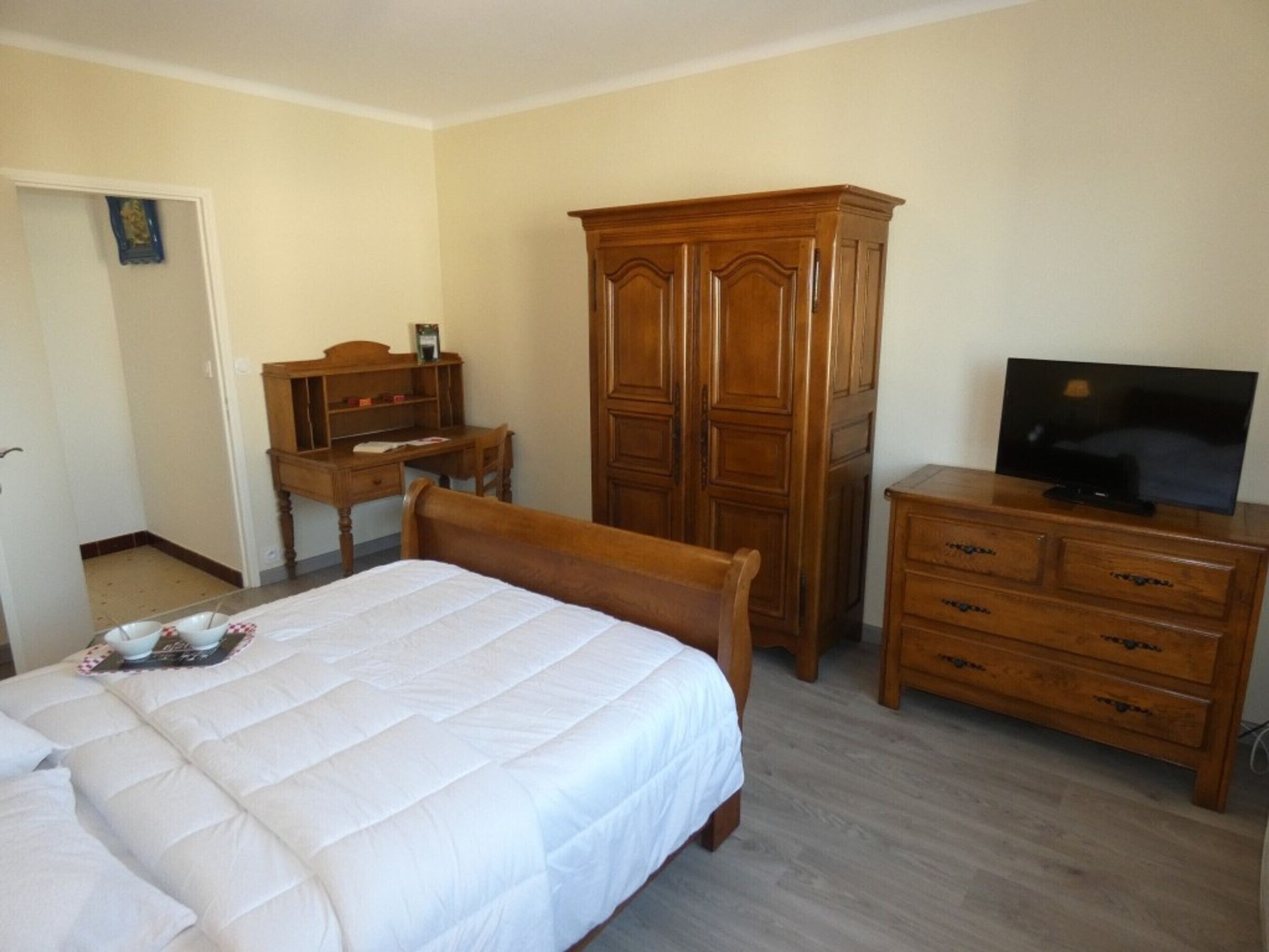 Un dormitorio con una cama, un armario de madera, un escritorio con silla y un televisor sobre una cómoda.