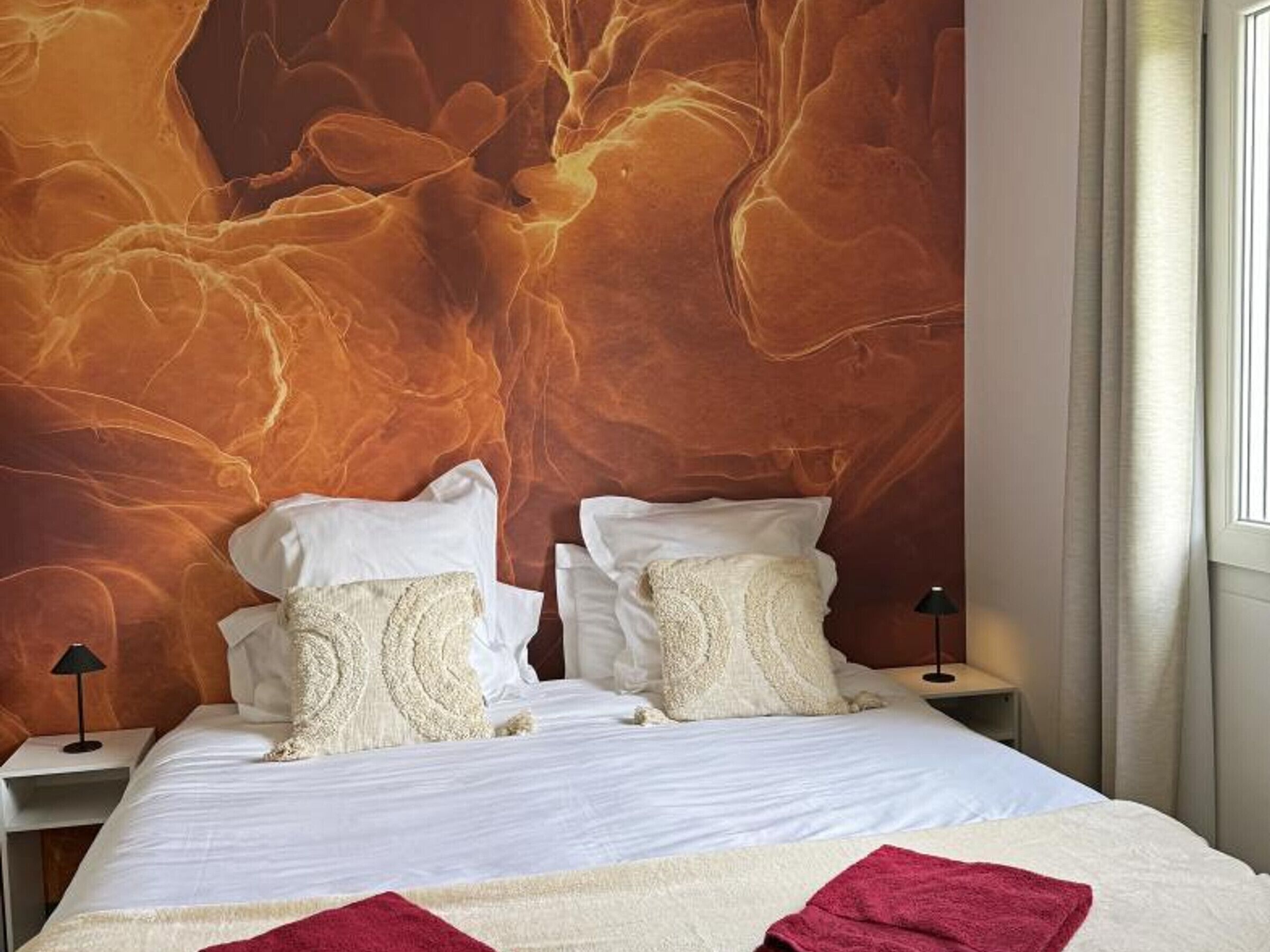 Una cama con sábanas blancas, dos almohadas, una manta roja y una pared con textura.