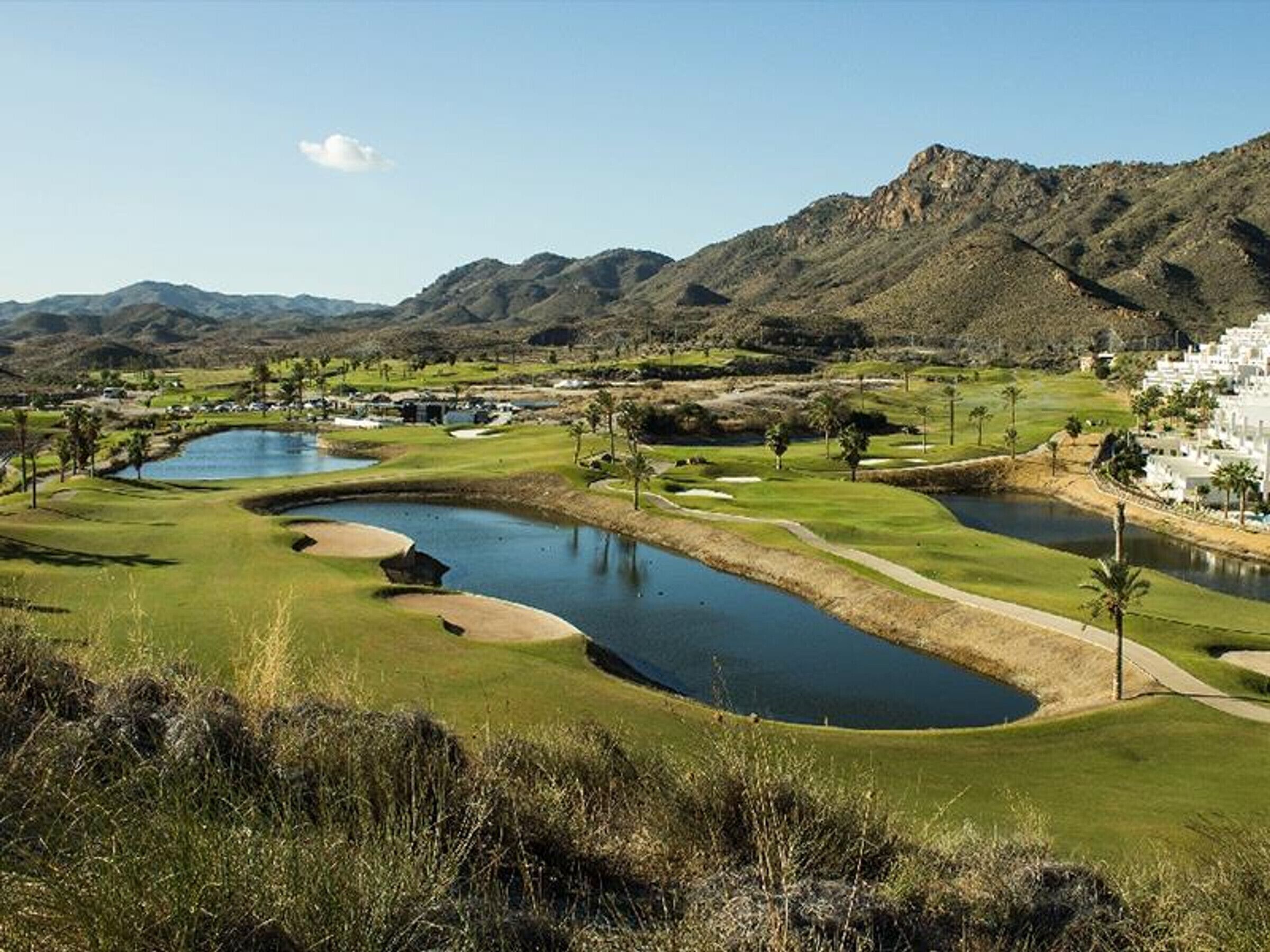 Um campo de golfe com lago, bunkers de areia e montanhas ao fundo.