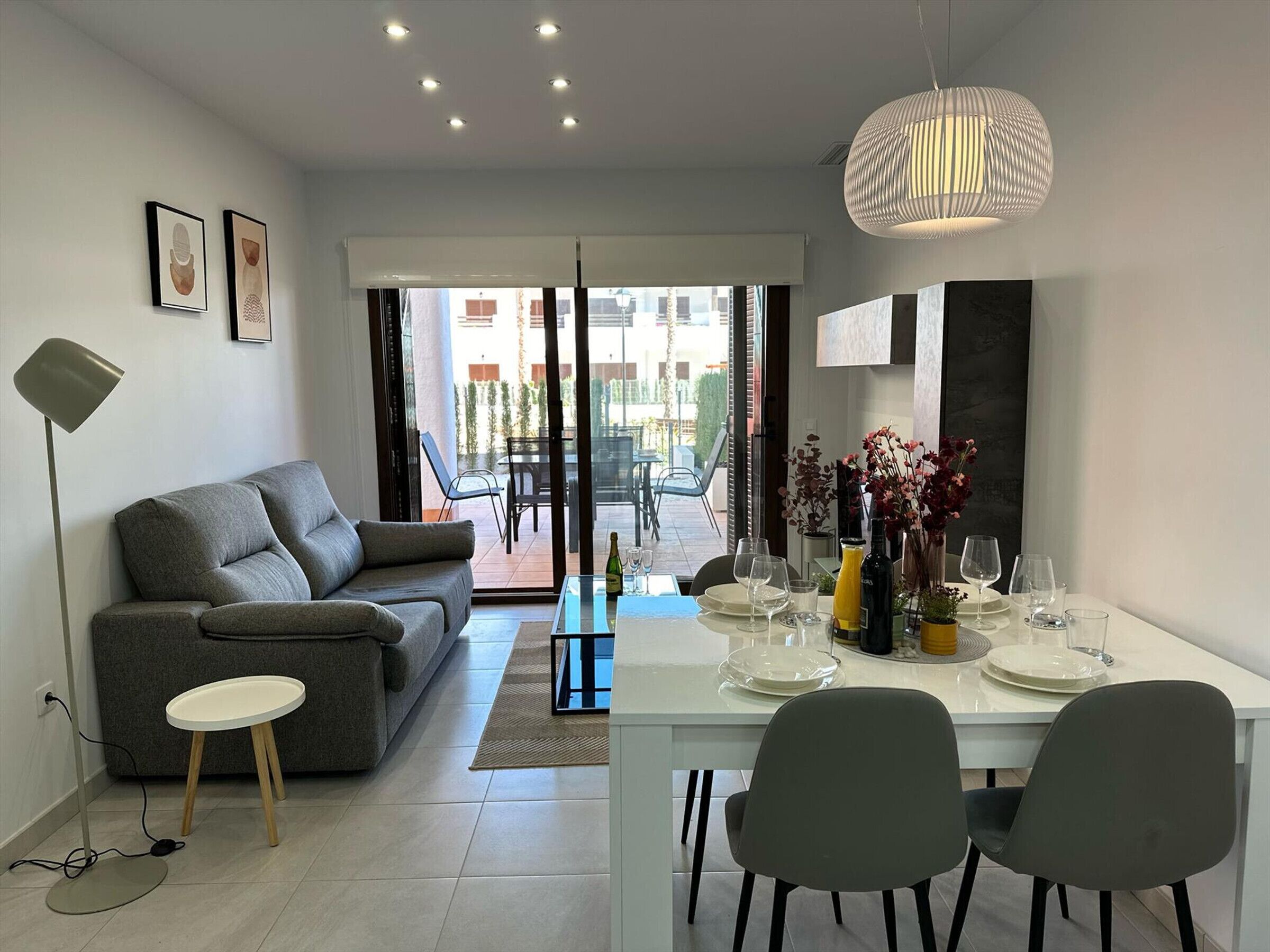 Uma sala moderna com um sofá cinzento, uma mesa de jantar posta para quatro pessoas e uma grande janela com vista para o exterior.