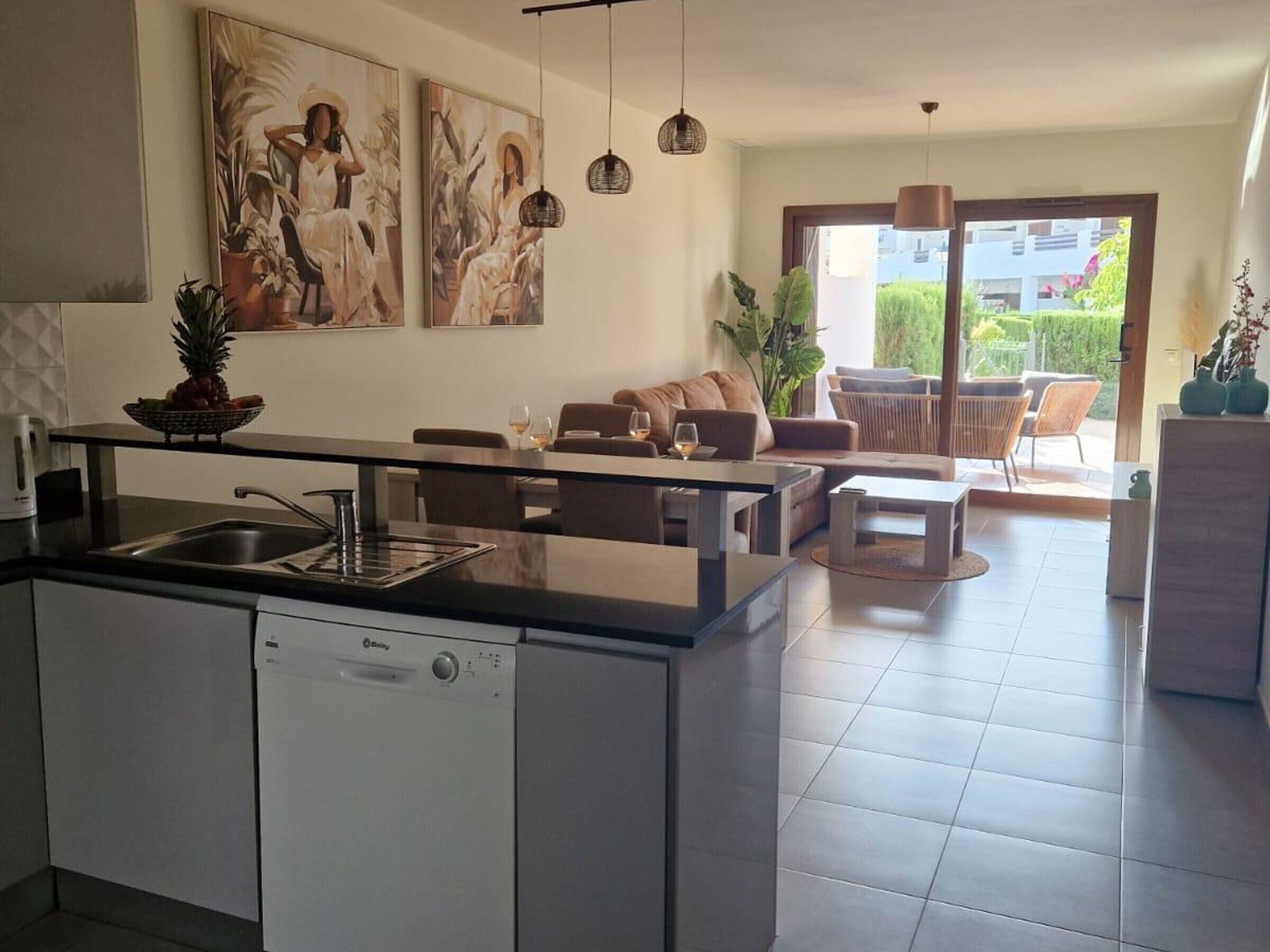 A modern kitchen with a dishwasher, a dining area with a table and chairs, and a living area with a sofa and a coffee table. There are two paintings on the wall and a plant on the windowsill.
