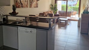 Una cocina moderna con lavavajillas, un comedor con mesa y sillas, y una sala de estar con sofá y mesa de centro. Hay dos cuadros en la pared y una planta en el alféizar de la ventana.