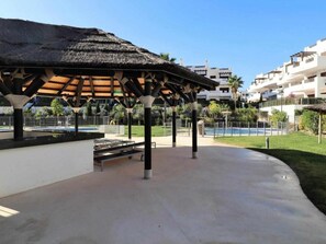 Un espacio exterior techado con techo de paja, una piscina y un edificio blanco al fondo.