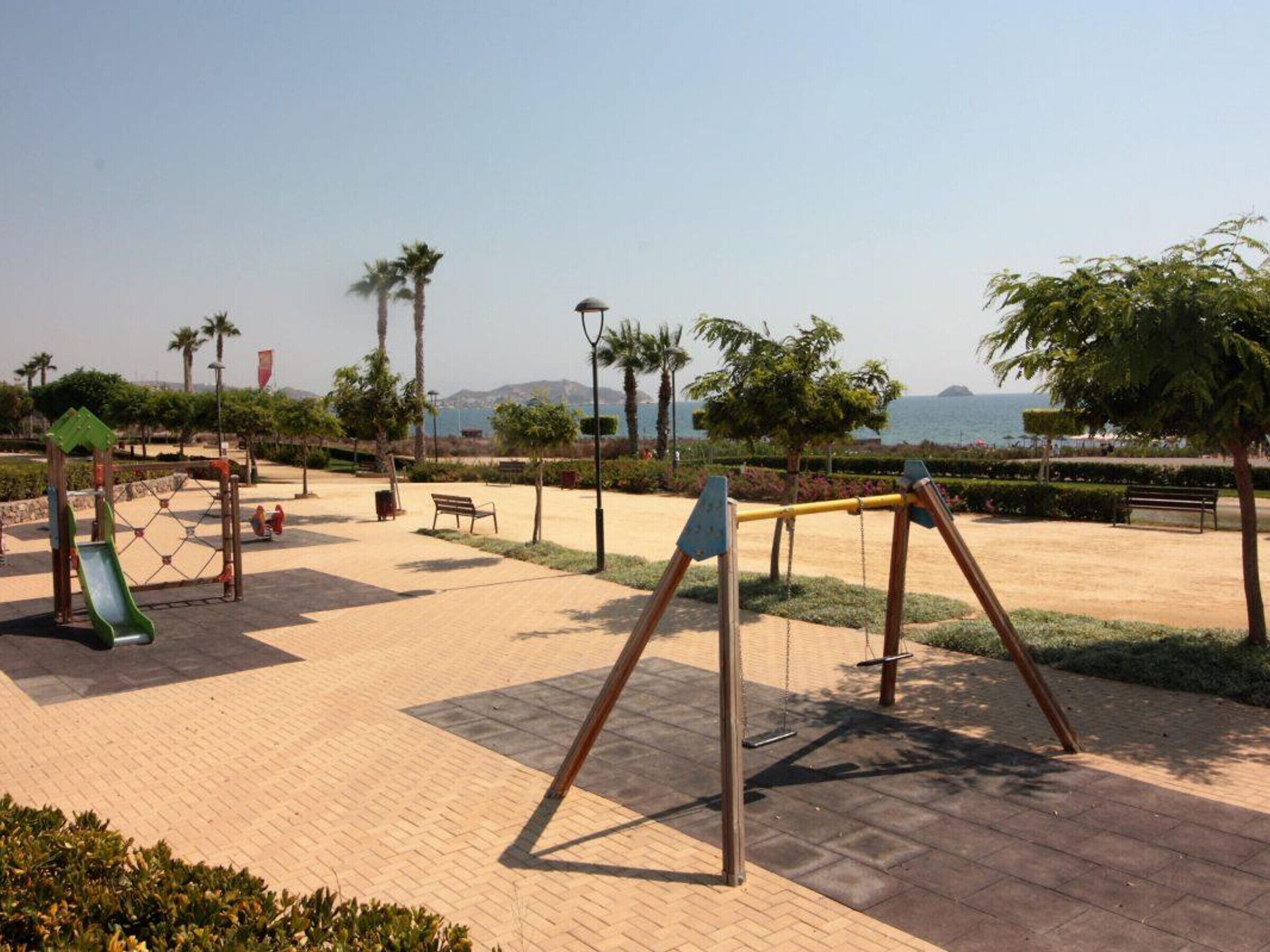 A playground with swings, a slide, and benches in a park with palm trees and a view of the sea.