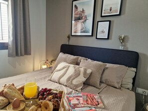 Ein ordentlich bezogenes Bett mit einer Zeitschrift, einem Tablett mit Frühstücksangeboten und einem Fenster mit Vorhängen.