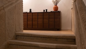 A wooden cabinet with a vase on top, placed in a hallway with a stone wall and a wooden floor. - Ando Living - São Miguel House - 203 (Porto)
