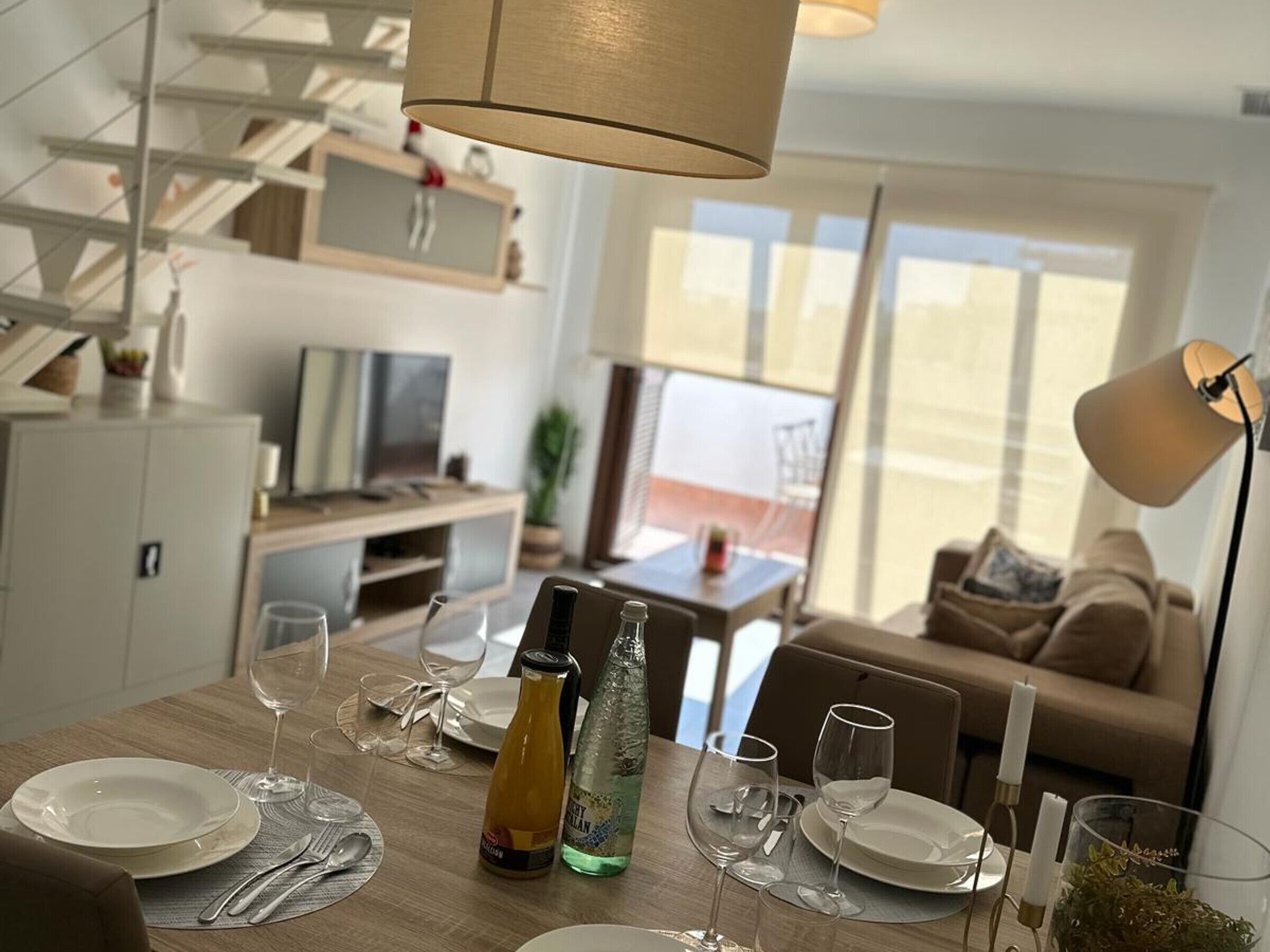 A modern dining area with a wooden table set for two, a bottle of water, and a bottle of wine. There is a sofa and a TV in the background.