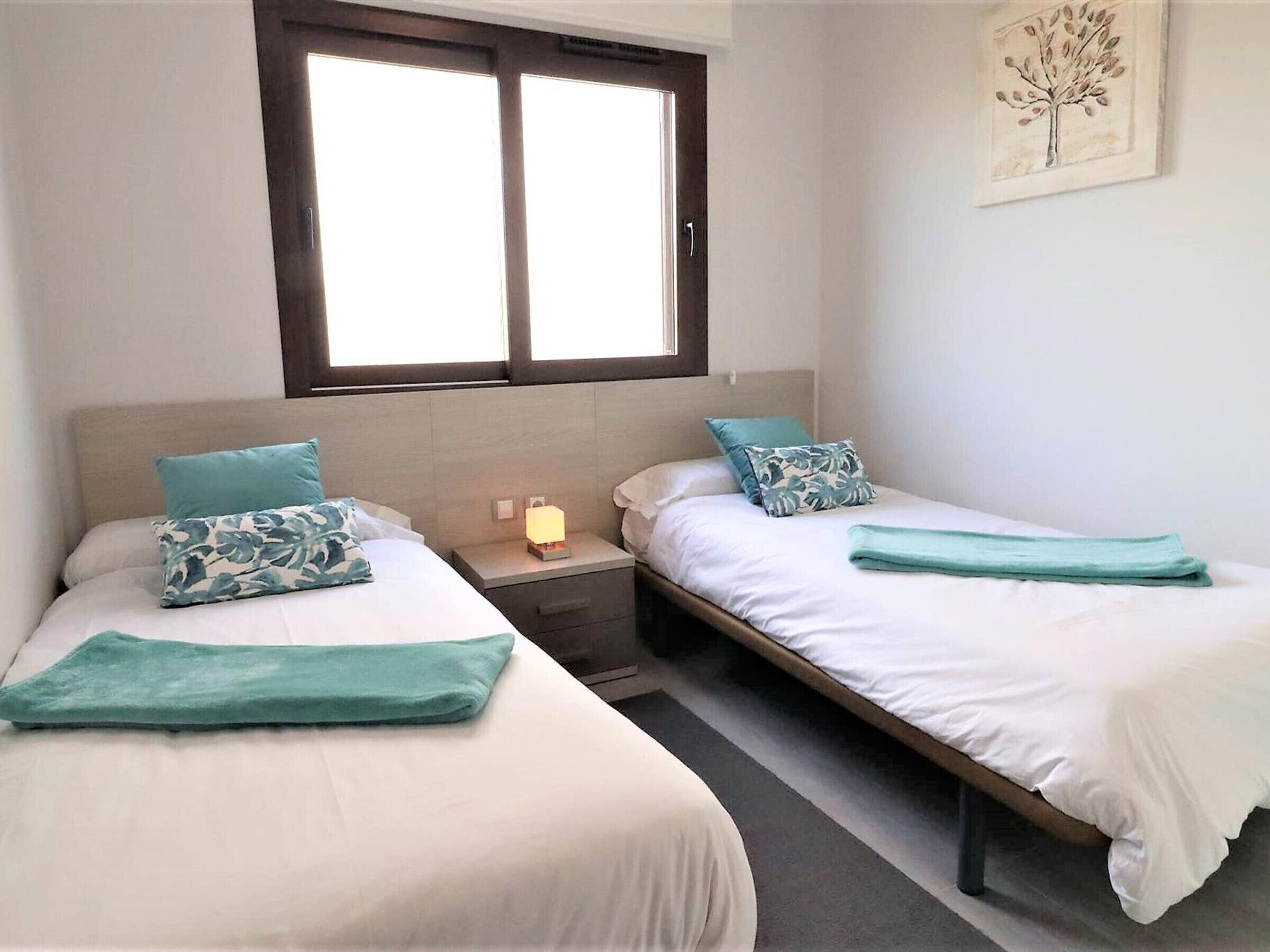 A room with two single beds, each with a teal throw blanket and matching pillows. There is a nightstand with a lamp between the beds.