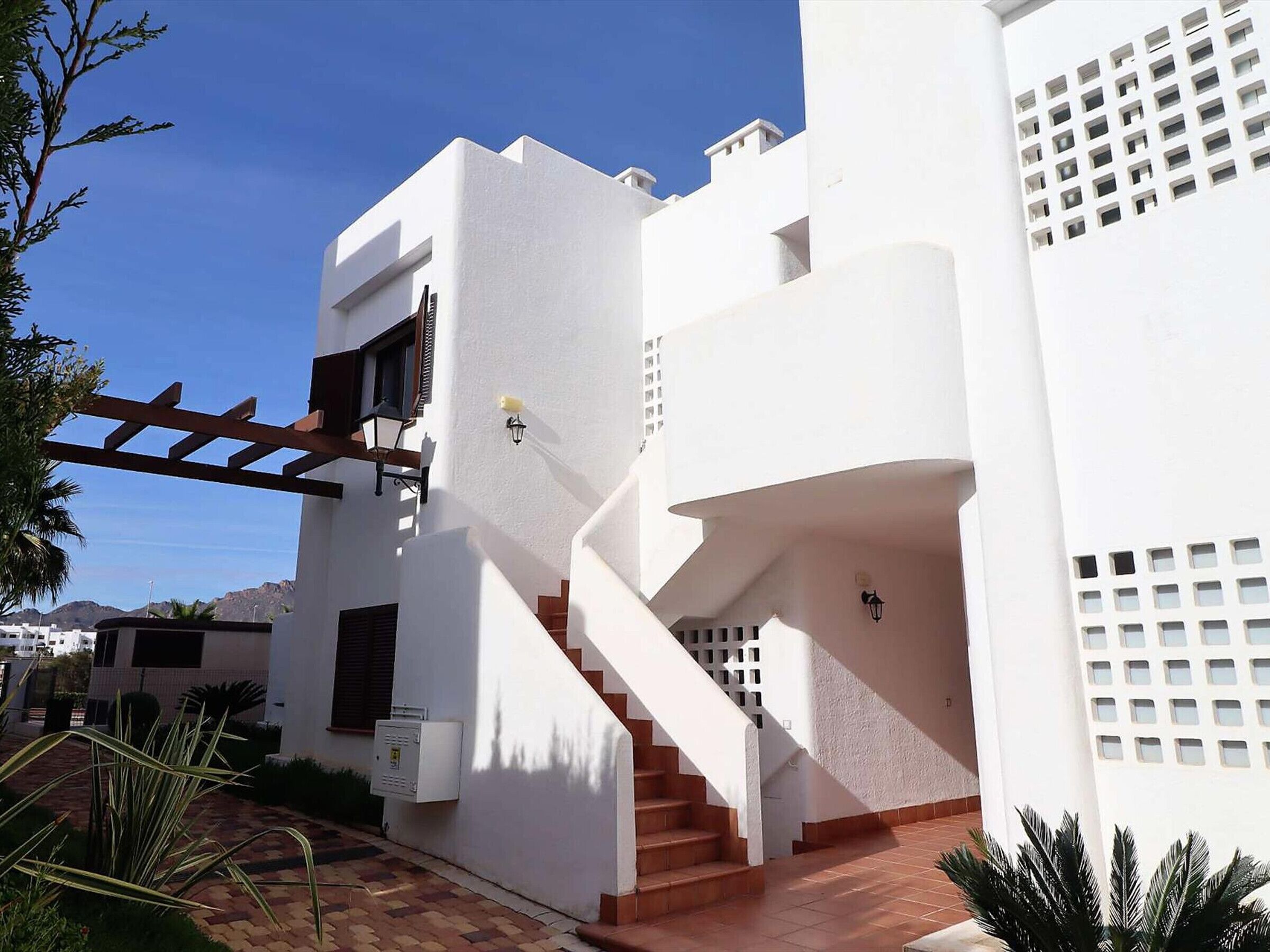 Un edificio blanco con una escalera que lleva al segundo piso, una pérgola y un pequeño espacio exterior con plantas.