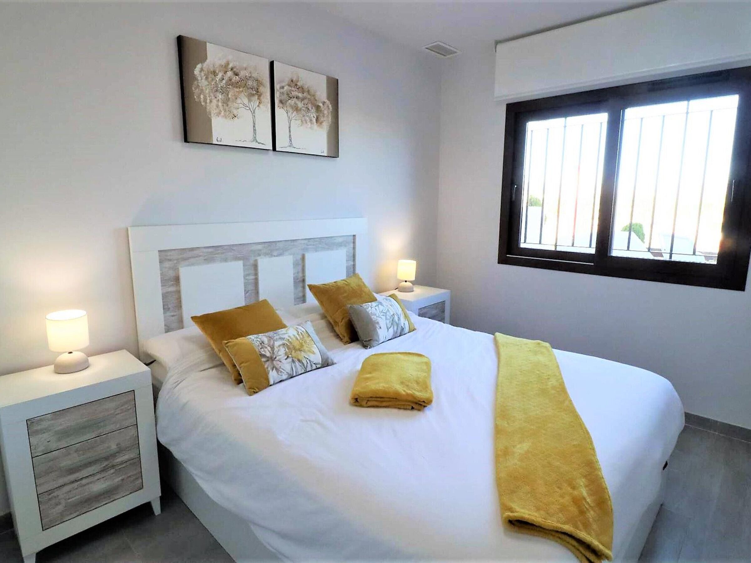 Une chambre à coucher bien rangée, avec un lit, deux tables de chevet, une fenêtre et deux tableaux accrochés au mur.
