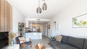 Una sala moderna con un sofá gris, una mesa de centro de madera y un televisor sobre un mueble blanco. Se observa un área de cocina al fondo, con una barra y lámparas colgantes.