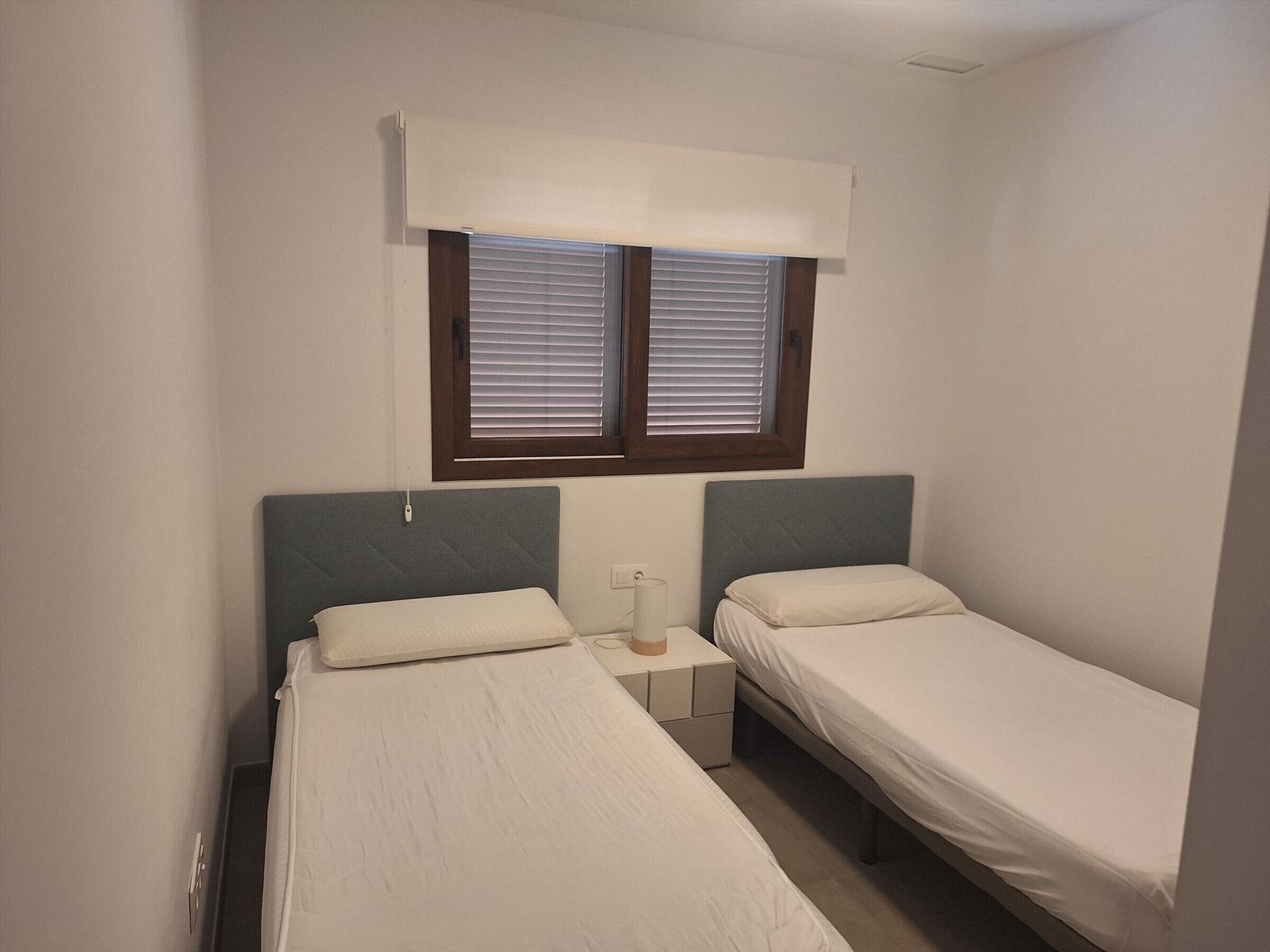 A small, single-room accommodation with two single beds, a bedside table with a lamp, and a window with blinds.