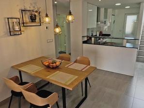 A modern dining area with a wooden table, chairs, and a fruit bowl. There is a kitchen visible through an open door in the background. - Charming apartment 200m from beach with communal garden (San Juan de los Terreros)