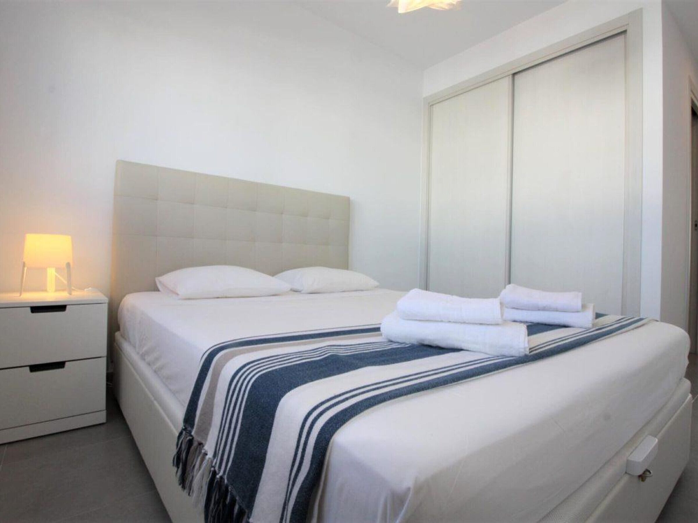 A neatly made bed with white linens and a striped blanket, a bedside lamp, and a closed wardrobe.