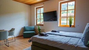 A bedroom with a bed, a TV mounted on the wall, a window with a view of greenery, a small table with chairs, and a potted plant. - Old schoolyard - Apartment Pausenhof (Oberstaufen)
