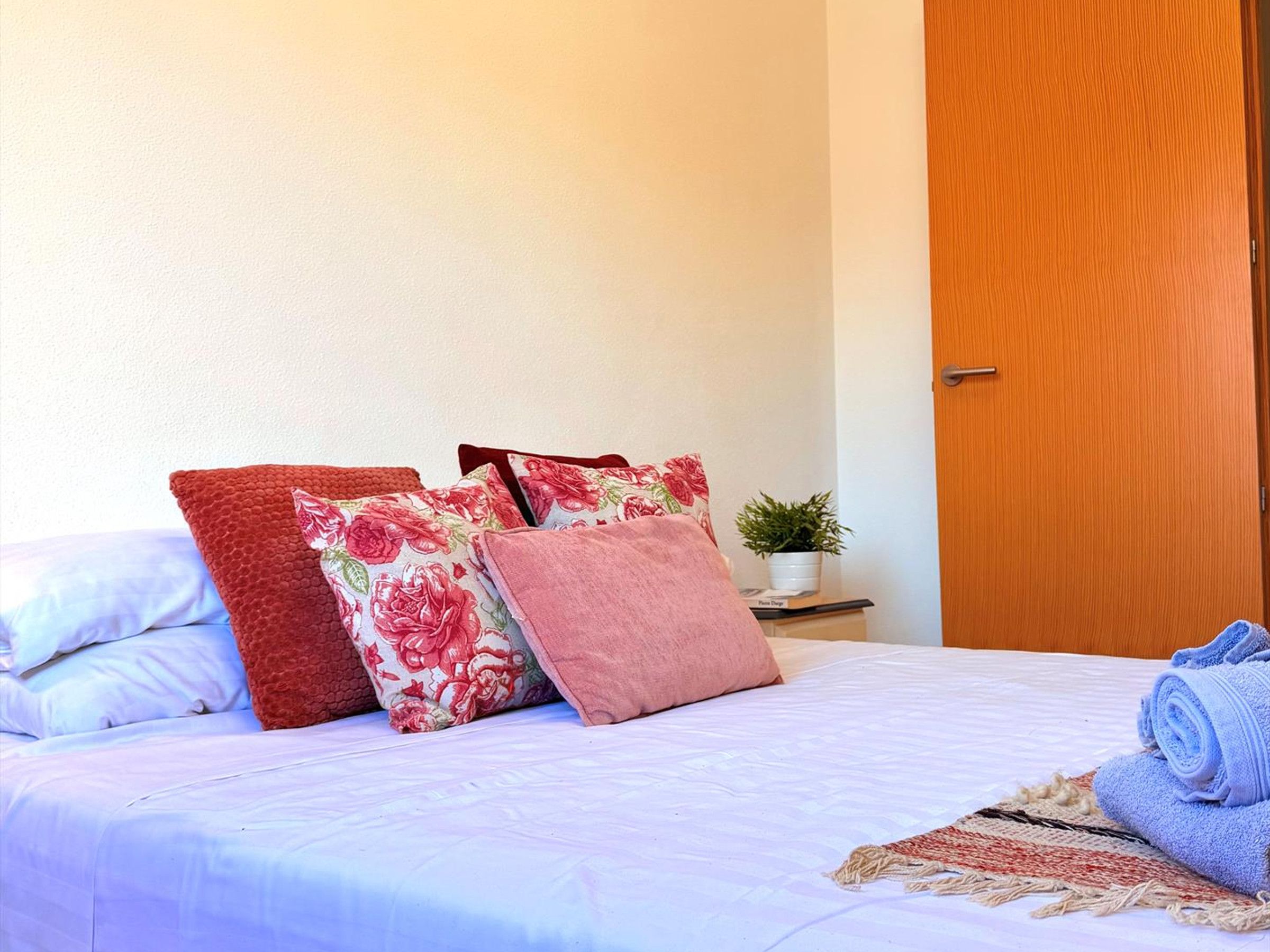 A neatly made bed with floral and solid-colored pillows, a small bedside table with a plant, and a wooden door in a room with plain walls.