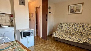Un espace de vie compact comprenant un coin cuisine, une table à manger avec de l’assiette, un four à micro-ondes, un canapé recouvert d’un tissu à motifs et une porte donnant sur une autre pièce.