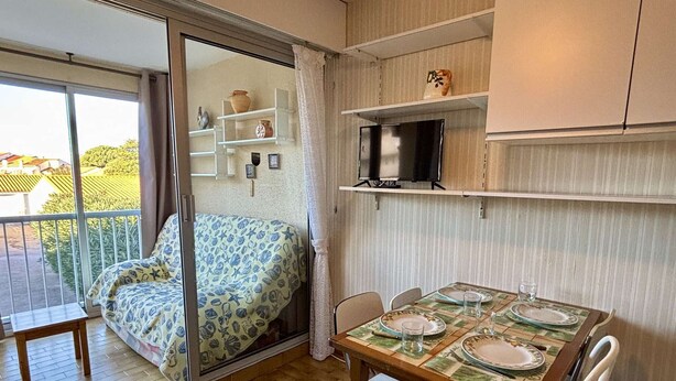 Une pièce confortable avec une porte-fenêtre coulissante donnant sur un balcon, une table à manger dressée avec des assiettes et des verres, et un téléviseur posé sur une étagère.