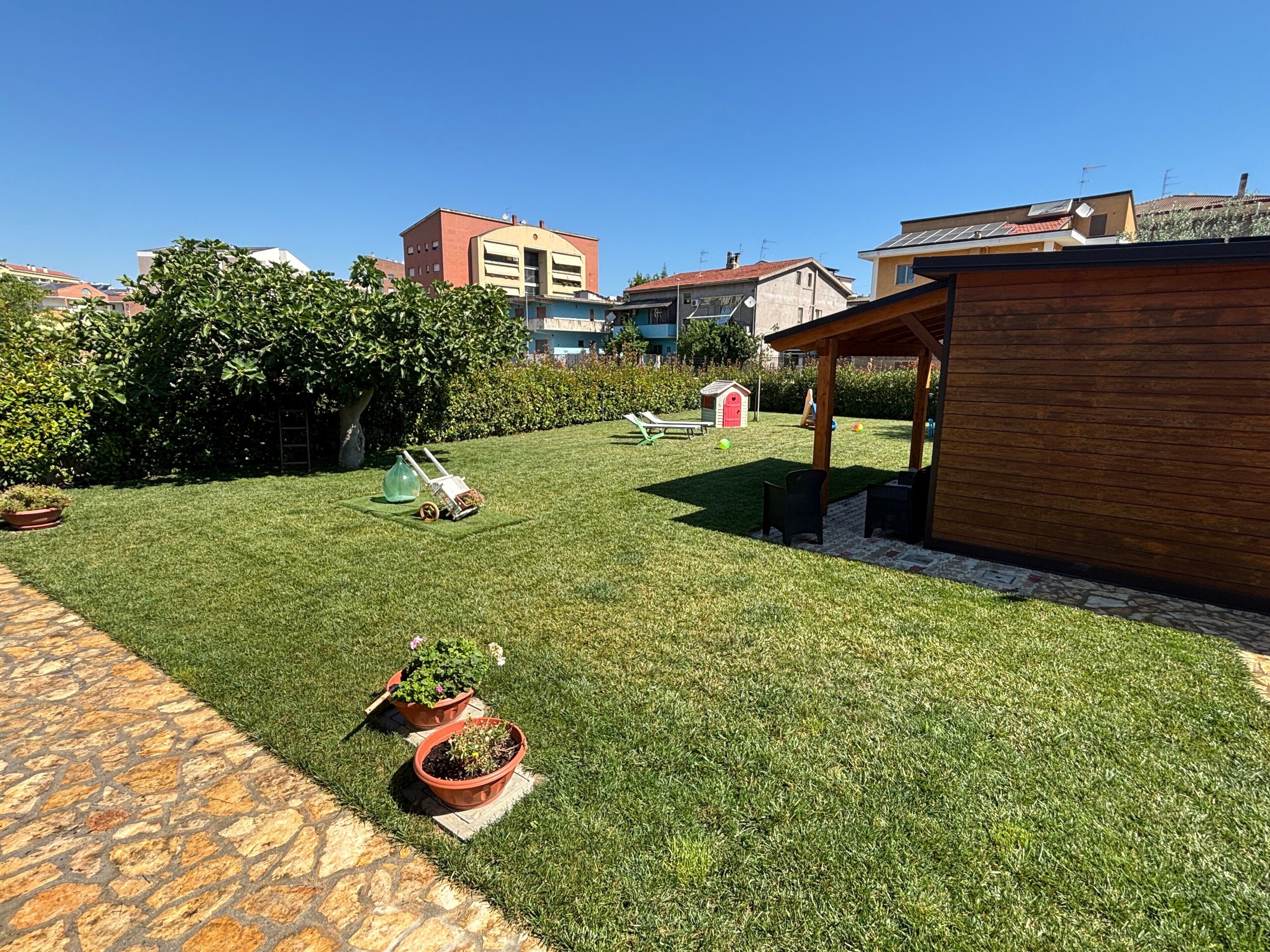Un prato curato con una pergola in legno, piante in vaso e un piccolo deposito rosso.