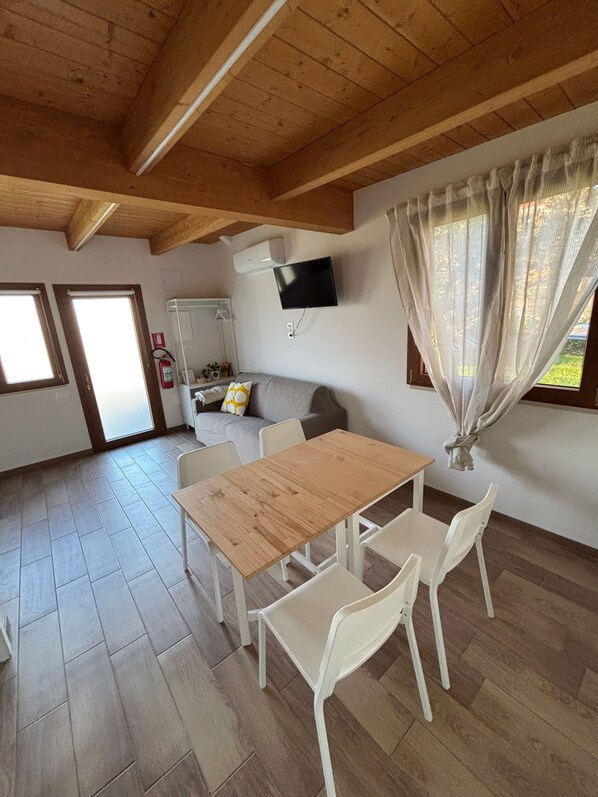 A room with a wooden ceiling, a TV mounted on the wall, a table with chairs, and a sofa.