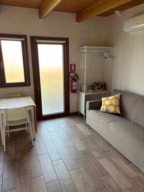 A room with a wooden floor, a white table with a chair, a sofa with a patterned cushion, a fire extinguisher, and a window with blinds.