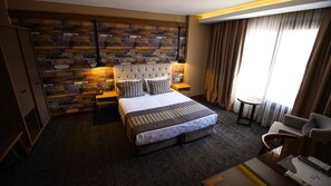 A hotel room with a large bed, a wooden accent wall, a nightstand, a lamp, a chair, and a window with curtains.