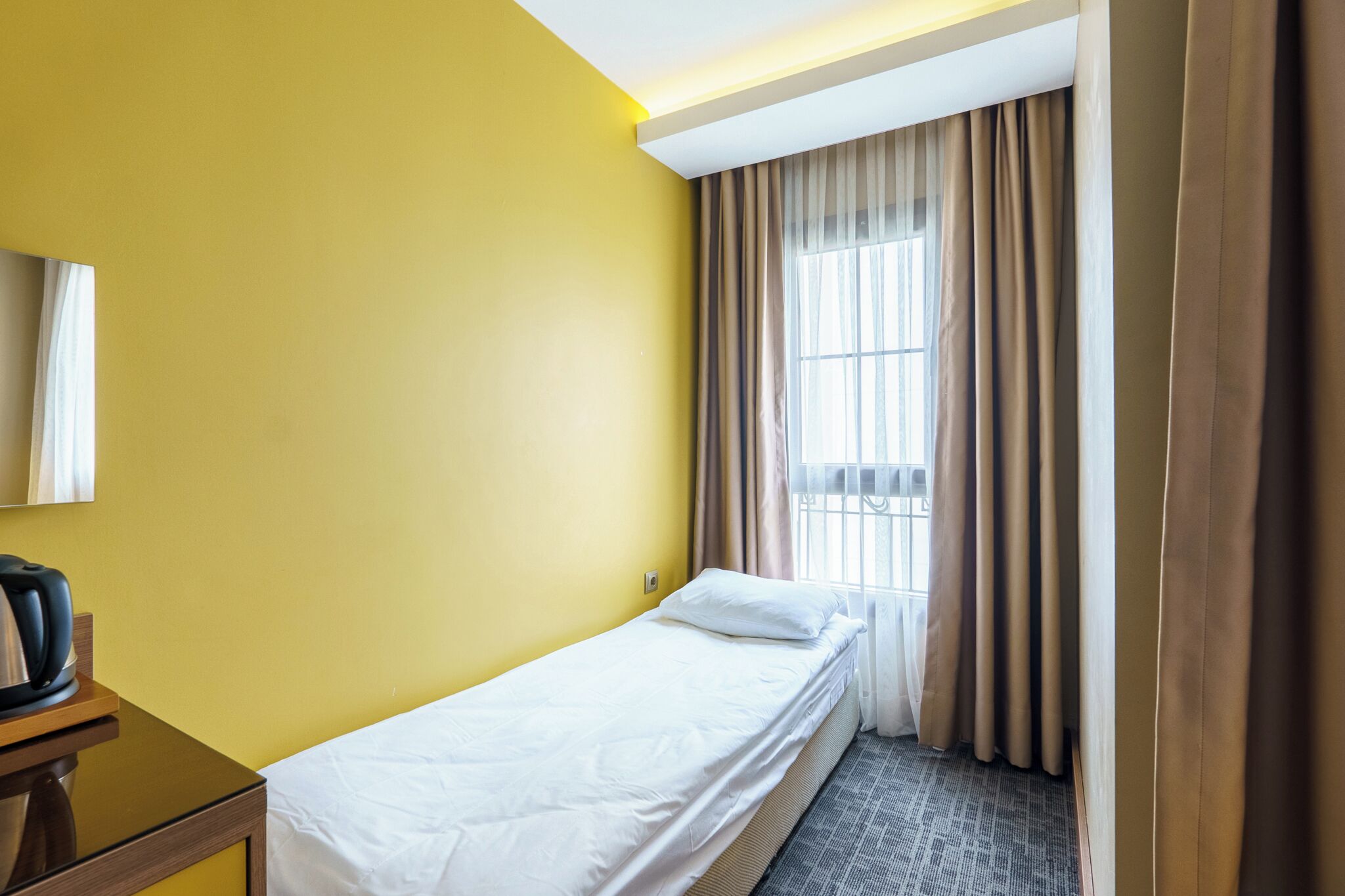 A small, single-bed room with a yellow wall, a window with curtains, and a bedside table with a kettle.