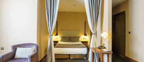 A hotel room with a bed, a chair, a desk with a lamp, and curtains.