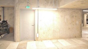 En korridor av betong med en dörr som är märkt med en skylt för nödutgång, en takmonterad lampa och en motorcykel som står parkerad till vänster.