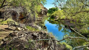 Paisaje natural con terreno rocoso, un banco y una masa de agua tranquila rodeada de vegetación.