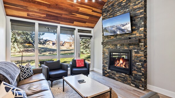 A modern living room with a stone fireplace, leather sofas, and large windows offering a view of the outdoors. - Prime Location: Modern Mountain Charm, Hot Tub, Sleeps 10, Close to Skiing & Dining! (Park City)