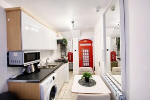 Une petite cuisine équipée d’un four à micro-ondes, d’un évier et d’un lave-linge, une porte de cabine téléphonique rouge et une petite table avec une plante.