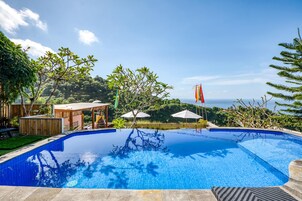 Una piscina azul cristalina con muelle de madera y sombrillas, rodeada de exuberante vegetación y vistas al océano.