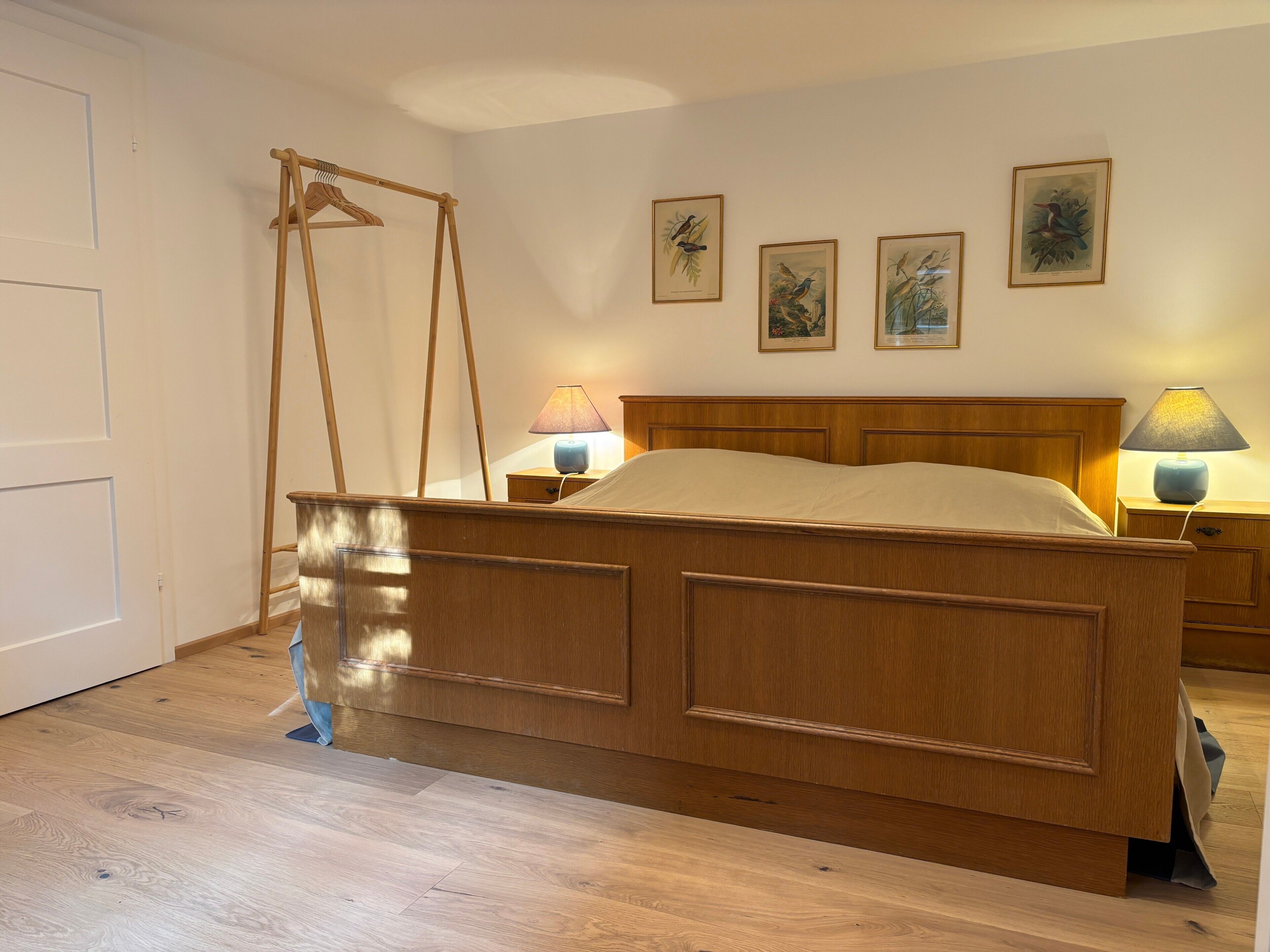 A bedroom with a wooden bed, two bedside lamps, a wooden wardrobe, and three framed wall art pieces.