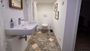 Ein Badezimmer mit weißem Waschbecken, WC und Duschvorhang. Der Boden ist mit gemusterten Fliesen belegt und an der Wand hängen zwei Bilder in Rahmen.
