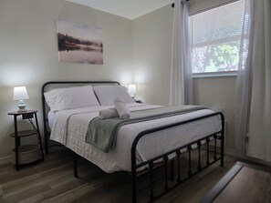 Un lit bien fait, avec une couette blanche et des oreillers, une table de chevet avec une lampe, une fenêtre avec des rideaux et un tableau accroché au mur.