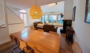 Ein großzügiger Essbereich mit einem Holztisch und Korbstühlen, eine Couch im Hintergrund und ein großes Fenster, das für natürliches Licht sorgt.
