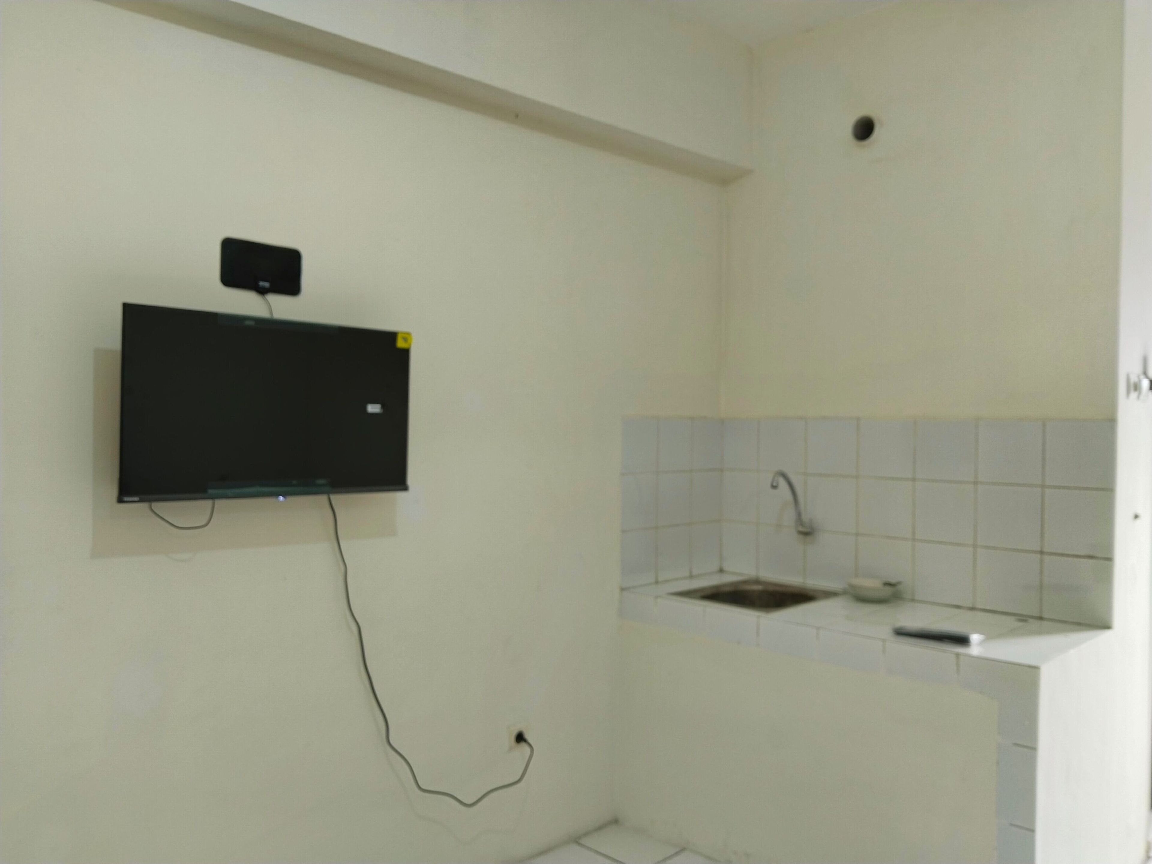 Un baño pequeño con paredes blancas, televisor montado, lavamanos y retrete.