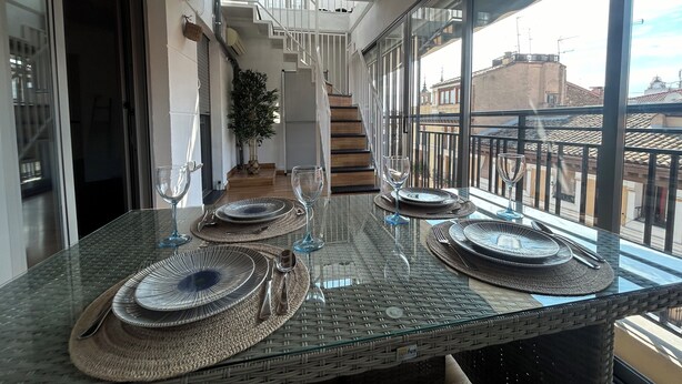 Un coin repas avec une table en verre dressée pour quatre personnes, un escalier et une vue sur le paysage urbain à travers de grandes fenêtres.